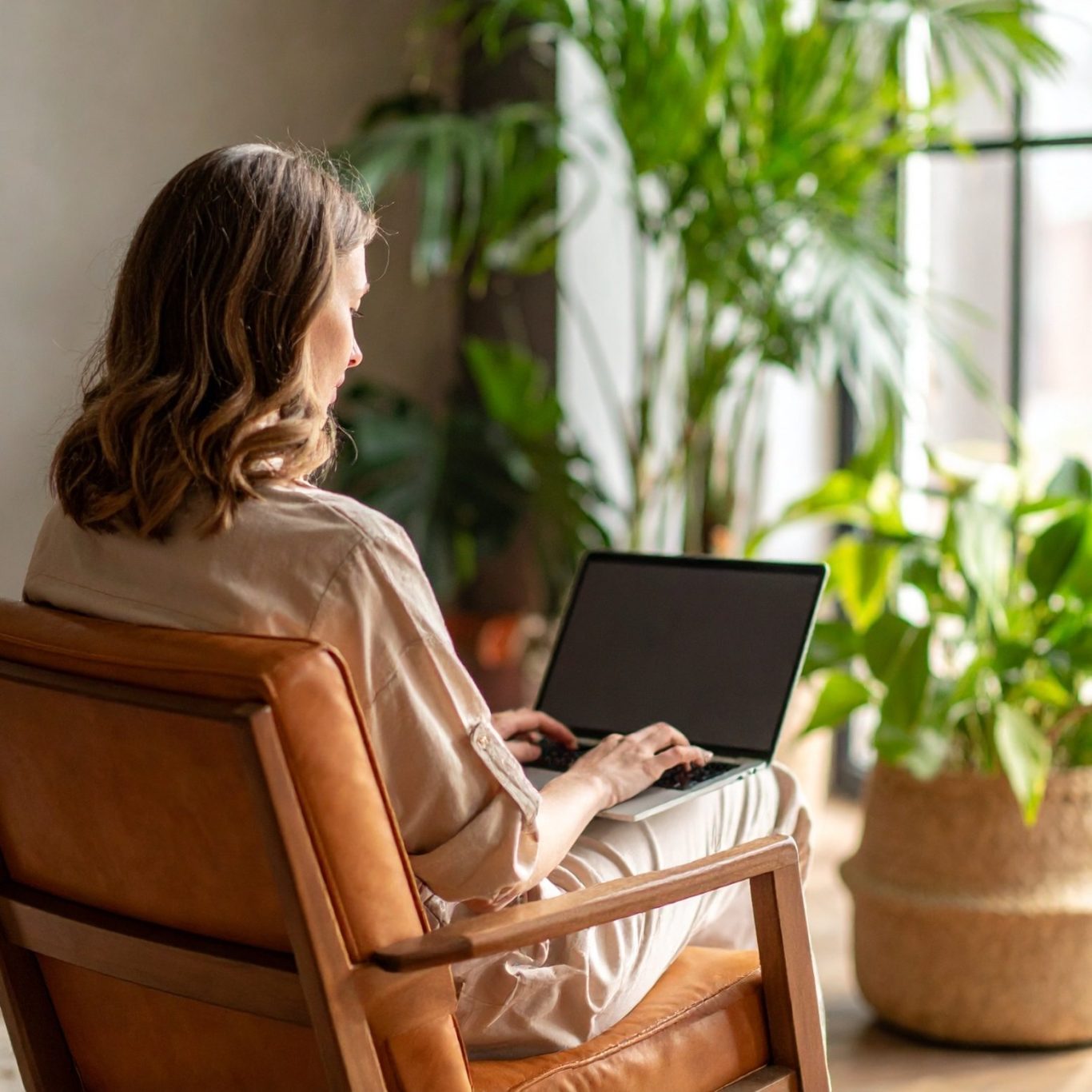 Frau mit Locken sitzt auf einem Sessel und arbeitet am Laptop, umgeben von Pflanzen.
